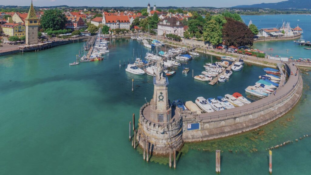 Vue sur le port de Lindau avec le phare et le lion de Bavière, les maisons à pignons de l’île et les sommets alpins en arrière-plan sous le ciel lumineux de mai.