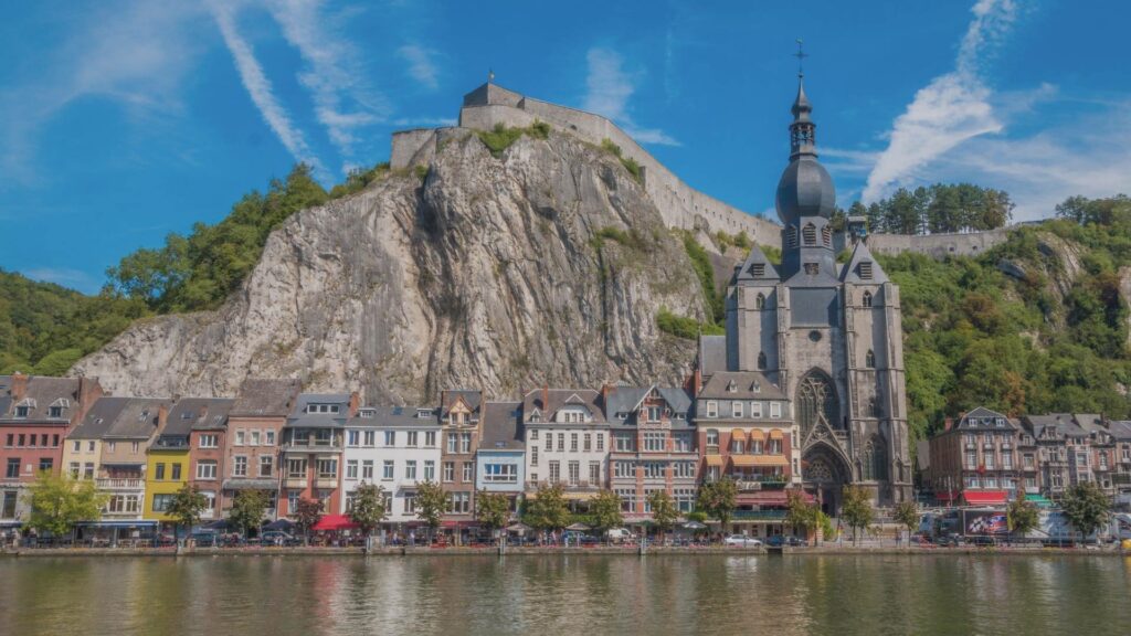 Vue de Dinant au bord de la Meuse avec la collégiale, les maisons colorées, la citadelle perchée et la falaise imposante sous un ciel doux de mars.