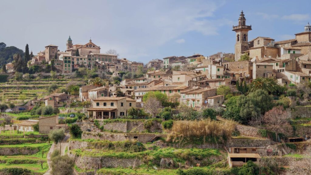 Vue printanière sur le village de Valldemossa avec ses maisons en pierre, ses cyprès et les montagnes de la Serra de Tramuntana en arrière-plan sous un ciel lumineux.