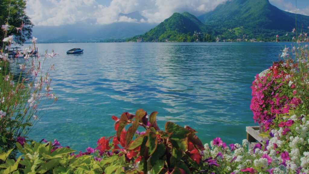 Vue sur les canaux fleuris d’Annecy avec les Alpes en arrière-plan et les eaux turquoise du lac scintillant sous le soleil de juillet.