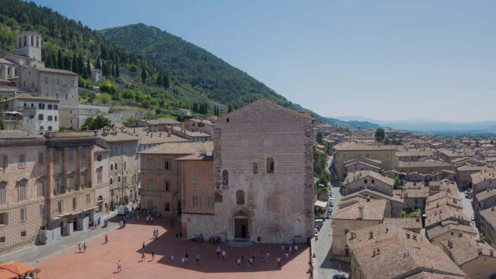 Vue panoramique sur Gubbio, avec ses toits en terre cuite et ses bâtiments en pierre grise, accrochés à flanc de colline sous un ciel légèrement voilé de printemps.
