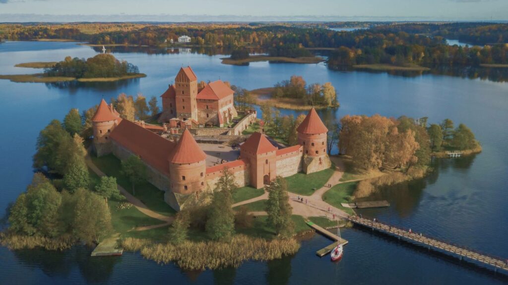 Vue panoramique sur le château de Trakai posé au centre du lac Galvė, relié par une passerelle en bois, sous un ciel bleu d’août.