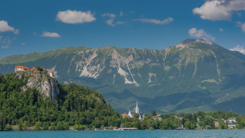 Vue panoramique sur le lac de Bled, son île avec l’église au centre, et le château perché sur la falaise, sous un ciel bleu de juillet.