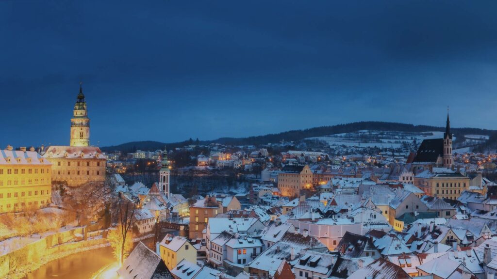 Vue hivernale de Český Krumlov avec ses toits enneigés, la rivière Vltava enserrant le centre-ville médiéval et le château dominant la scène, dans une atmosphère calme et brumeuse.